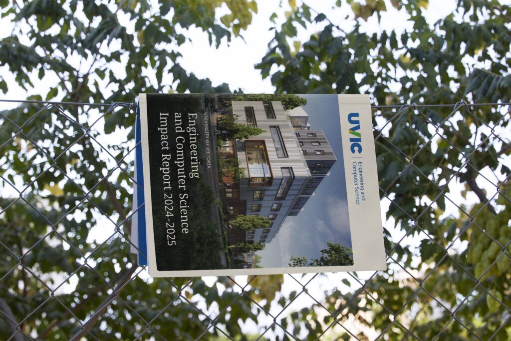 Printed copy of the University of Victoria Faculty of Engineering and Computer Science Impact Report displayed on a chain-link fence, showcasing Steady Studio's editorial design against a backdrop of sunlit trees.